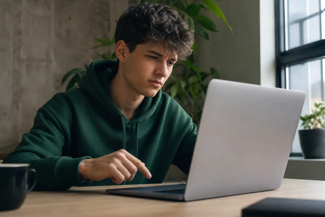 Jeune personne assise à un bureau, ordinateur portable ouvert, doigt suspendu au-dessus du pavé tactile comme hésitant avant de cliquer