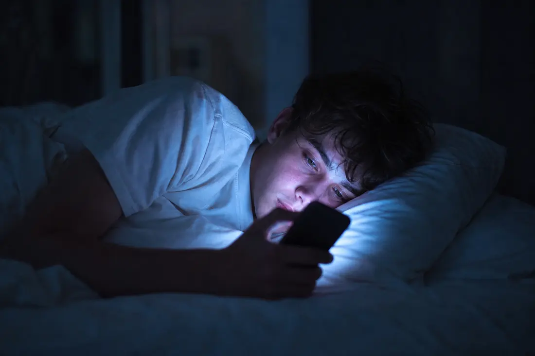 Jeune allongé dans son lit la nuit, éclairé par la lumière bleutée de l'écran de son téléphone en train de faire défiler un fil d'actualité