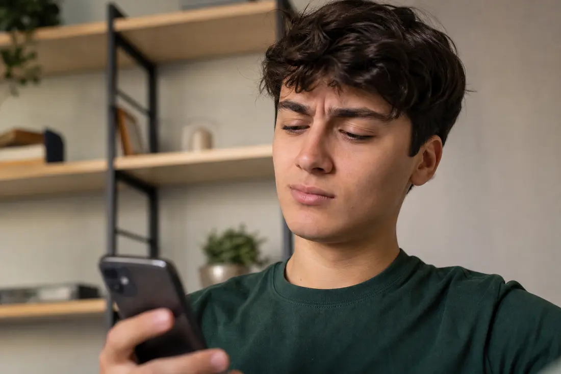 Jeune homme en tee-shirt vert, l'air sceptique, regardant l'écran de son téléphone dans un salon aux étagères en bois
