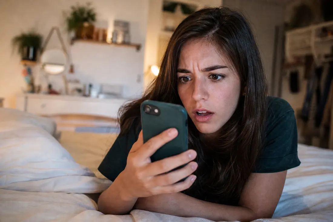Jeune allongé sur son lit qui regarde son téléphone avec une expression surprise en voyant une publicité