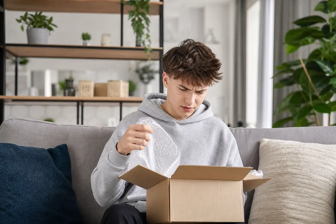Un jeune homme en sweat gris, assis sur un canapé, sort du papier bulle d'un petit carton avec une mine dubitative
