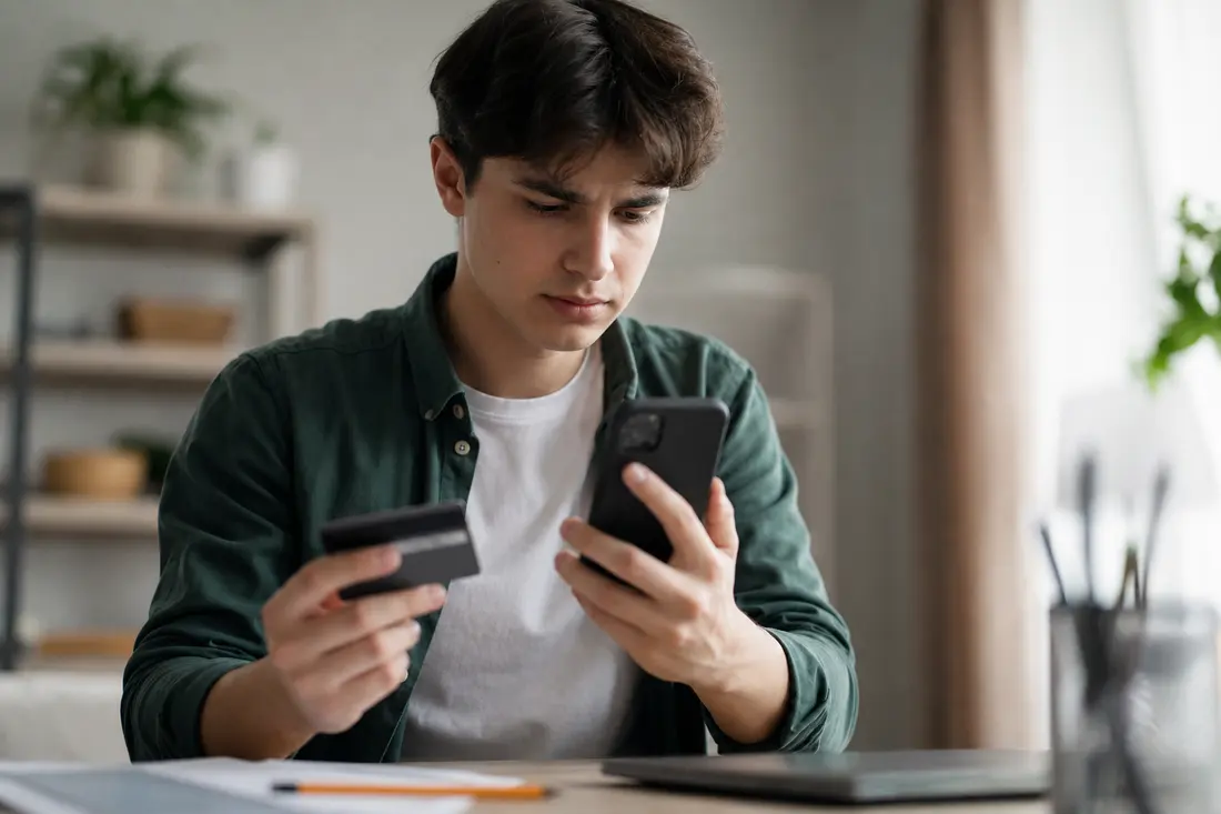 Jeune personne regardant son application bancaire sur smartphone, carte bancaire posée à côté, expression concentrée