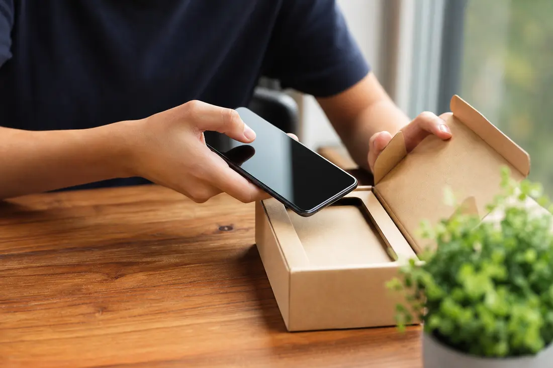 Mains d'un jeune tenant un smartphone déballé d'une boîte en carton neutre posée sur une table en bois