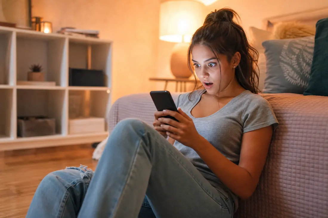 Jeune femme assise par terre contre son lit, smartphone dans les mains, expression de surprise en découvrant quelque chose à l'écran