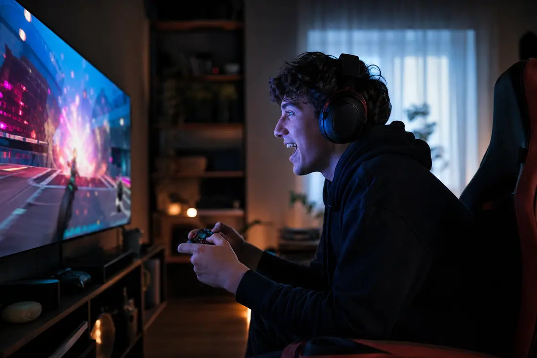 Jeune devant son écran de jeu vidéo, manette en main, en train d'ouvrir un coffre virtuel qui projette une lumière colorée