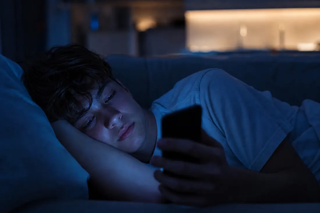 Jeune allongé sur un canapé le soir, éclairé par la lumière bleutée de son téléphone en train de scroller