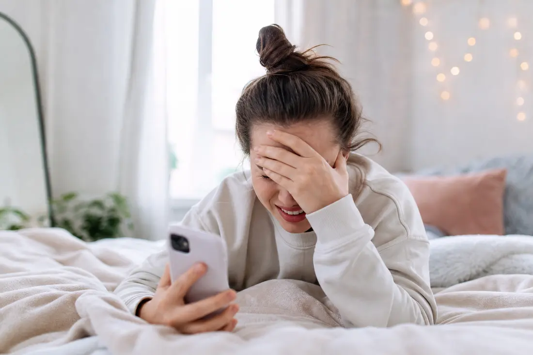 Jeune qui se cache le visage avec la main en regardant son téléphone, expression gênée, dans une ambiance lumineuse et colorée