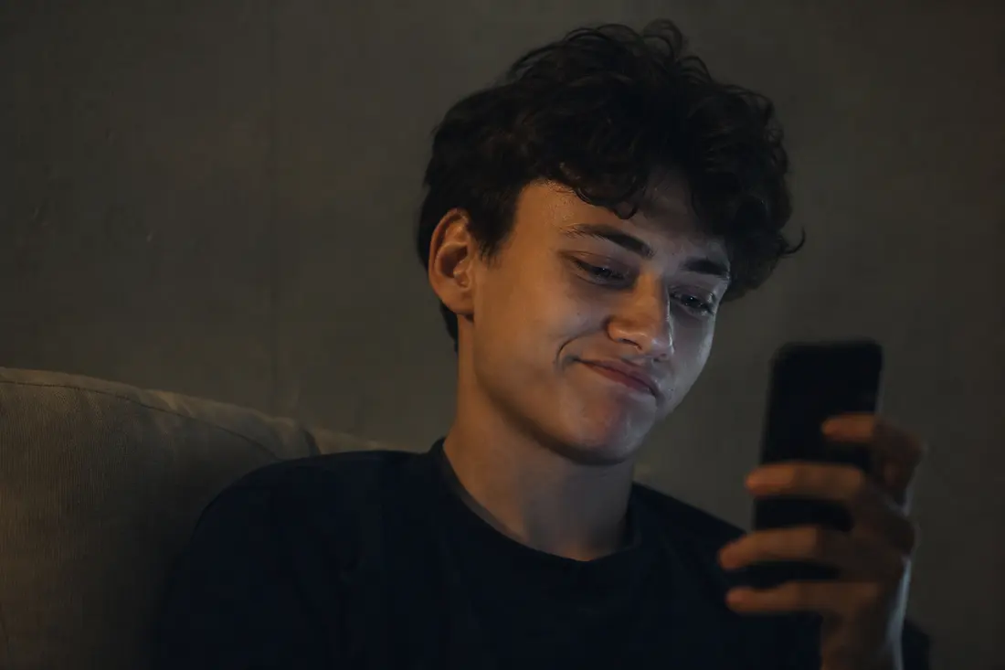 Jeune personne souriant avec une expression ambiguë en regardant son téléphone, éclairage contrasté
