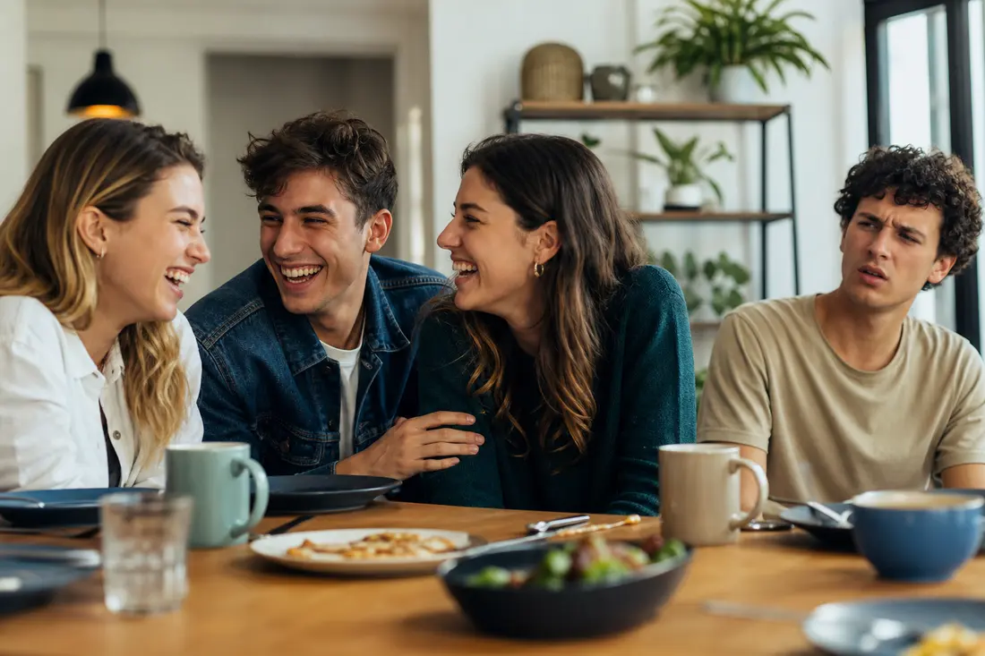 Groupe d'amis riant ensemble autour d'une table, complices, pendant qu'une autre personne à côté reste sans réaction