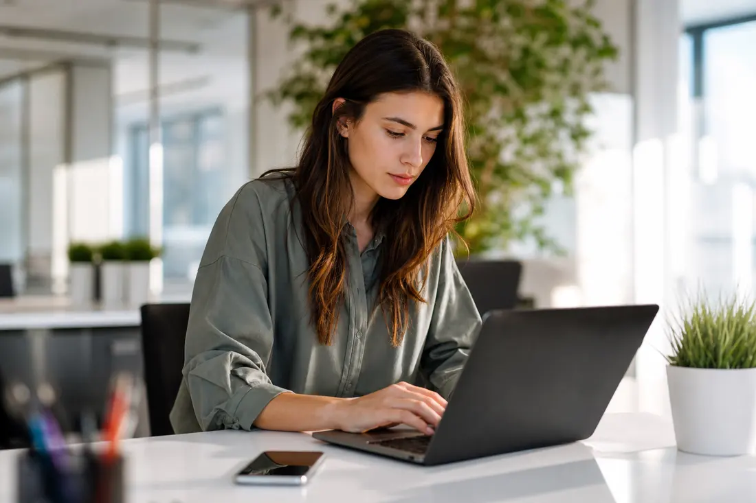 Jeune community manager travaillant sur un ordinateur portable avec un smartphone à côté, bureau lumineux et moderne