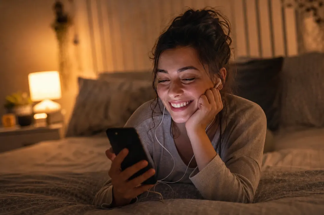 Jeune femme souriante allongée sur son lit, regardant une vidéo sur son smartphone avec des écouteurs, ambiance chambre chaleureuse le soir