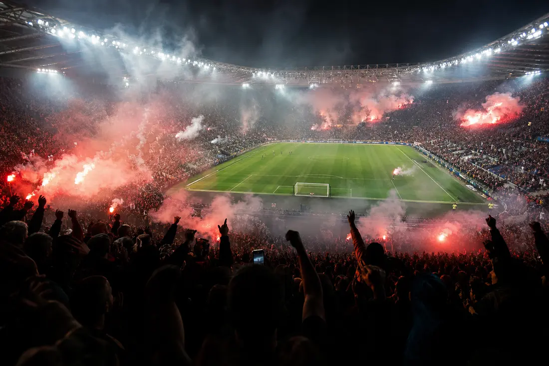Ambiance de stade de foot rempli avec fumigènes et supporters en tribune lors d'un match sous les projecteurs