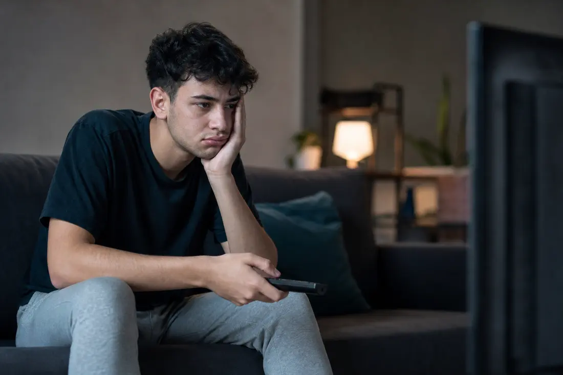 Jeune déçu assis dans son canapé devant la télévision, télécommande à la main, en train de lire une notification d'annulation de série