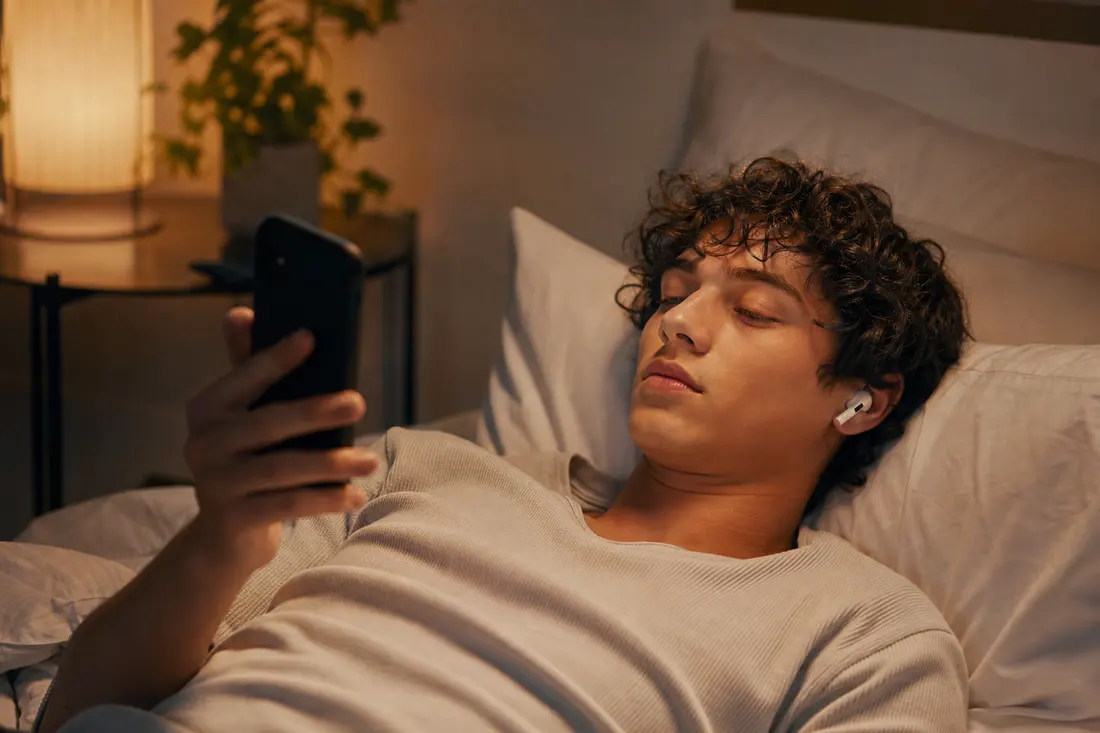 Jeune allongé sur son lit, écouteurs sur les oreilles, écoutant de la musique sur son téléphone dans une chambre éclairée