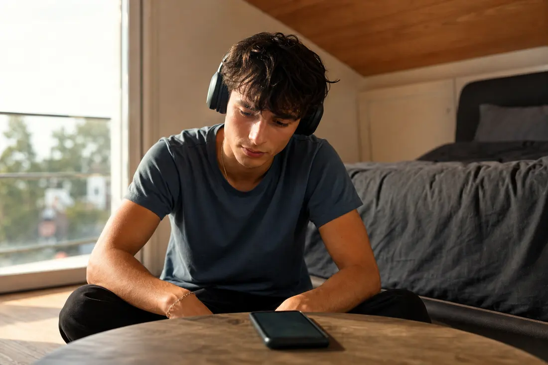 Jeune homme avec un casque audio sur les oreilles dans une chambre, regardant son téléphone posé devant lui, lumière douce de fin de journée